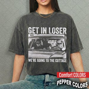 May include: A charcoal grey mineral-washed t-shirt featuring a white graphic of two people in a car wearing sunglasses. The text above reads GET IN LOSER and the text below reads WE'RE GOING TO THE COTTAGE. The shirt has short sleeves and a crew neck.