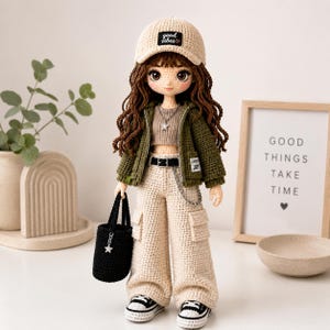 Op de afbeelding: Een handgemaakte gehaakte pop met lang bruin haar, gekleed in een beige honkbalpet met een zwarte patch met de tekst good vibes, een olijfgroen vest met rits, een beige crop top, een beige cargobroek met een zwarte riem en zwart-witte sneakers.