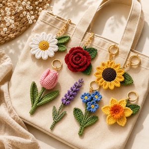 Può includere: Una collezione di portachiavi a fiori all'uncinetto fatti a mano, tra cui una margherita, una rosa, un tulipano, lavanda, nontiscordardimé, un girasole e un narciso. Ogni portachiavi ha un anello color oro ed è attaccato a una borsa tote beige.