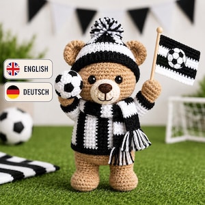 May include: A tan crocheted teddy bear wearing a black and white striped jumper, a matching scarf with fringe, and a beanie with a pom-pom. The bear holds a small crocheted football in one hand and a black and white flag with a football in the other.