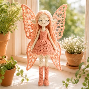 May include: A crochet doll featuring a peach-colored dress and matching boots, with long peach yarn hair and large, intricate, lace-like wings in a matching peach hue. The doll has a cream-colored face with closed eyes and pointed ears.