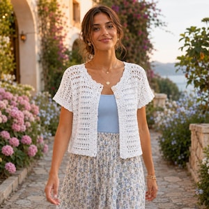 May include: A white short-sleeved crochet cardigan featuring an open-knit pattern, a scalloped neckline, and two visible buttons at the front. The garment is worn over a light blue ribbed tank top and a floral print skirt with a tiered design.