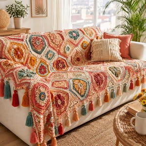 May include: A colorful, crocheted throw blanket with a geometric pattern covers a white sofa. The blanket features a variety of colors, including peach, coral, teal, and yellow. Tassels in coordinating colors hang from the edge of the blanket. Decorative pillows complete the look.