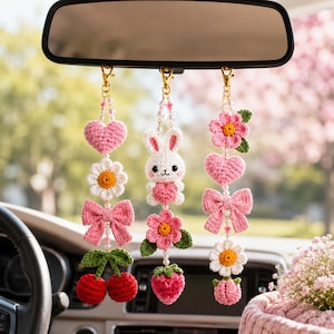 May include: Crocheted car charms hanging from a rearview mirror. The charms include a pink heart, daisy, bow, and cherries. Other charms include a white bunny holding a pink heart, a pink flower, and a strawberry. The charms are attached with gold-colored clips.
