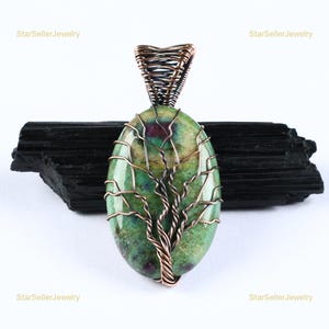 May include: An oval-shaped pendant featuring a green and magenta stone wrapped in dark copper wire. The wire is intricately twisted to form a tree design on the front of the stone, extending upwards to create a secure bail for hanging the pendant.