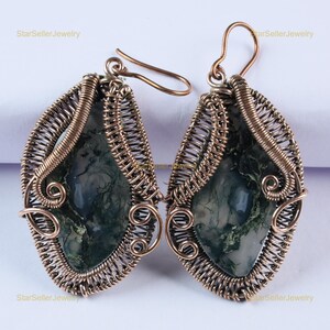 May include: A pair of dangle earrings featuring teardrop-shaped moss agate stones encased in intricate copper wire wrapping. The stones display dark green, moss-like inclusions against a translucent grey background, suspended from copper ear hooks.