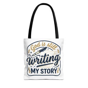 Pode incluir: Uma bolsa tote branca com alças pretas apresentando um design gráfico com o texto God is still writing MY STORY em azul marinho e dourado. A ilustração inclui uma caneta-tinteiro e uma folha de papel com pequenos detalhes de estrelas.