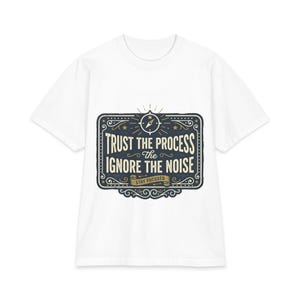 May include: A white short-sleeve t-shirt featuring a dark blue rectangular graphic with decorative borders. The design includes a compass icon and the text "TRUST THE PROCESS" and "IGNORE THE NOISE" in a cream-colored serif font, with "STAY FOCUSED" on a banner.