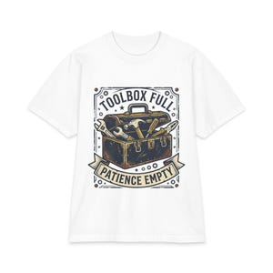 May include: A white short-sleeve t-shirt featuring a graphic illustration of a dark brown metal toolbox filled with various hand tools including a hammer, wrench, and pliers. The text reads TOOLBOX FULL above and PATIENCE EMPTY below the box.