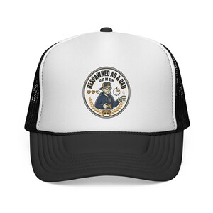 May include: A white and black trucker hat featuring a circular graphic on the front. The design shows a cartoon person with glasses holding a game controller and a mug, with the text "RESPAWNED AS A DAD GAMER" and three heart icons.