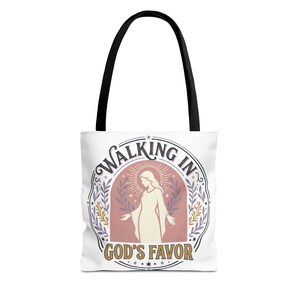 Può includere: Una borsa tote bianca con manici neri che presenta una grafica centrale di una figura in un arco con il testo WALKING IN GOD'S FAVOR. Il design include un sole, stelle e rami in tonalità tenui di rosa, viola e oro.