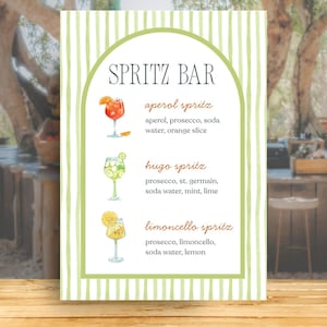 Op de afbeelding: Een rechthoekig bord met een lichtgroene gestreepte rand en een boogvormig kader. De tekst luidt SPRITZ BAR, met de ingrediënten voor een Aperol spritz, Hugo spritz en Limoncello spritz, elk vergezeld van een aquarelillustratie van het drankje.