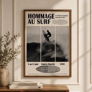 May include: A framed black and white poster with the text "HOMMAGE AU SURF" and "La Première Exposition Française de Surf". The poster features a surfer riding a wave. Additional text includes "3 au 8 Aout", "Galerie Biarritz", and "1989".