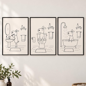 May include: A set of three framed wall art prints featuring black line drawings on a cream background. The illustrations depict a cartoon dog performing bathroom routines: brushing teeth at a sink, sitting on a toilet, and bathing in a tub.