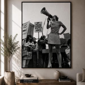 Könnte beinhalten: Ein schwarz-weiß gerahmter Wandkunstdruck, der eine Protestszene zeigt. Eine Person hält im Vordergrund ein Megafon, während andere Schilder mit der Aufschrift "US" und "UP FROM UNDER WOMEN UNITE" vor einer Kulisse aus Stadtgebäuden halten.