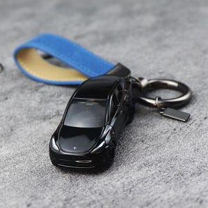 May include: A black die-cast model car keychain with a blue and tan leather strap. The keychain features a silver metal ring and a small rectangular tag. The car model has the Tesla logo on the hood.