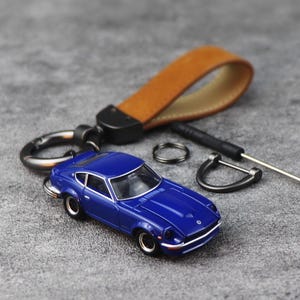 May include: A blue die-cast model car keychain with a brown leather strap. The keychain includes a keyring, a small screwdriver, and a black carabiner clip. The car is a classic sports car design.