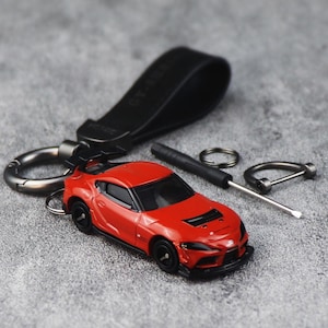 May include: A red sports car model keychain featuring a black rear spoiler, black wheels, and black trim. The keychain includes a black leather strap, a metal D-ring, a small circular ring, and a miniature flathead screwdriver for assembly.