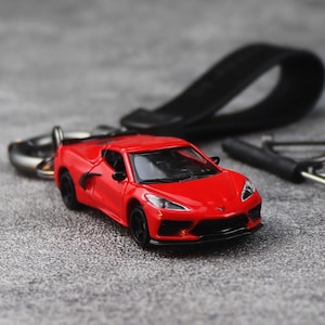 May include: A red miniature sports car keyring with black wheels and a black leather strap. The car is a detailed replica, showcasing the sleek design of a modern sports car. The keyring is on a grey surface.