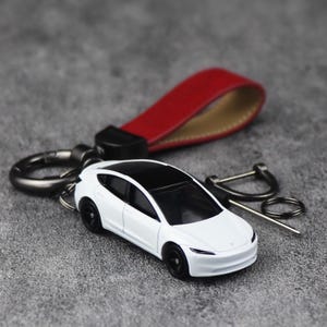 Op de afbeelding: Een witte speelgoedauto met een zwart dak en zwarte wielen is bevestigd aan een sleutelhanger met een rode leren band. De sleutelhanger heeft een zilveren sluiting en een zilveren ring. De auto is een miniatuurmodel.