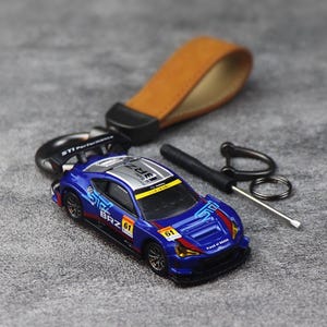 Op de afbeelding: Een blauwe Subaru BRZ speelgoedauto sleutelhanger met een beige leren band, een zwarte metalen ring en een kleine schroevendraaier. De auto heeft racestickers, het nummer 61 en STI Performance branding. Het dak is zilver met zwarte BRZ-tekst.