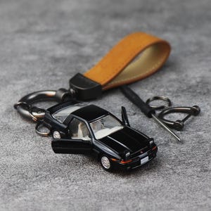 May include: A black miniature sports car keyring with open doors, featuring a tan leather strap, a dark metal carabiner, and a small screwdriver tool. The car has pop-up headlights, a detailed interior, and silver wheels on a dark grey surface.