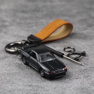 Könnte beinhalten: Ein schwarzer Modellauto-Schlüsselanhänger mit einem braunen Lederriemen und schwarzer Hardware. Das Auto hat silberne Felgen und rote Rücklichter. Auf einer grauen Oberfläche.