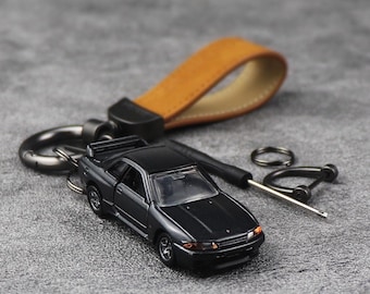 Può includere: Un portachiavi con un'auto modello nera, un cinturino in pelle marrone e hardware nero. L'auto ha cerchi argentati e fanali posteriori rossi. Su una superficie grigia.
