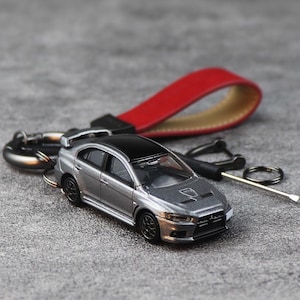 Pode incluir: Chaveiro de carro em miniatura prateado e preto com uma alça de couro vermelha. O carro tem um spoiler e detalhes. O chaveiro inclui um fecho de metal e um porta-chaves, para entusiastas.