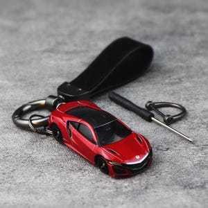 Può includere: Un portachiavi a forma di auto sportiva Honda NSX rossa con cinturino in pelle nera, moschettone in metallo scuro e un piccolo cacciavite. Il veicolo in miniatura presenta un tetto nero, ruote nere e l'emblema Honda sul cofano anteriore.