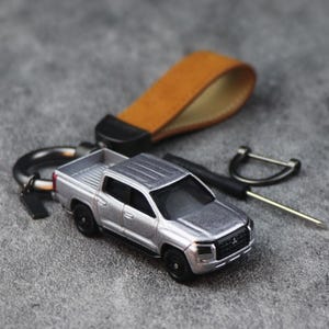 May include: A silver pickup truck keyring featuring a detailed miniature vehicle model with black wheels and a Mitsubishi logo on the front grille. The keyring includes a tan leather loop, a dark metal D-ring, and a small screwdriver tool.