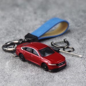 Könnte beinhalten: Ein Miniatur-Limousinenmodell in Rot, befestigt an einem blauen Lederschlaufen-Schlüsselanhänger. Der Schlüsselanhänger verfügt über einen dunklen Metall-Drehverschluss und einen Ring, ergänzt durch einen kleinen Metallschraubendreher.