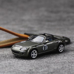May include: A metallic dark gray miniature convertible sports car featuring a black roll bar, silver five-spoke wheels, and white racing decals including the number 01 on the doors, a checkered pattern on the rear fenders, and Potenza text on the front.