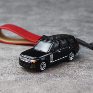 以下が含まれることがあります： 赤いレザーのキーホルダー用ループに取り付けられた、黒いレンジローバーSUVのミニチュアモデル。このおもちゃの車両は、シルバーカラーのホイール、レンジローバーのロゴが入った詳細なフロントグリル、黒いルーフを備えており、機能的なアクセサリーとしてデザインされています。
