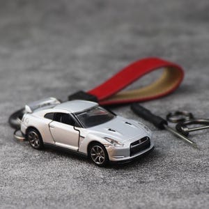 Op de afbeelding: Een zilveren modelauto-sleutelhanger met een rode leren band. De miniatuurauto heeft gedetailleerde kenmerken, waaronder wielen en een spoiler. De sleutelhanger bevat ook een kleine schroevendraaier en sleutelringen.