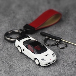 Könnte beinhalten: Ein weißer Sportwagen-Schlüsselanhänger mit schwarzem Dach, rotem Innenraum und weißen Rädern. Das Auto ist an einem dunklen Metallring und einem roten Lederband befestigt. Ein kleines schwarzes Werkzeug mit Metallstift ist enthalten.