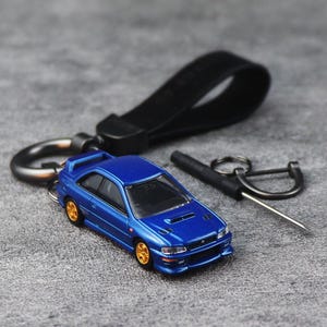 Könnte beinhalten: Ein blauer Miniatur-Auto-Schlüsselanhänger mit goldenen Rädern und einem Heckspoiler. Der Schlüsselanhänger umfasst ein schwarzes Lederband, einen D-Ring aus Metall und ein kleines Schraubendreher-Werkzeug. Das Modell hat klare Fenster und Details.