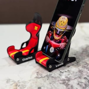 Op de afbeelding: Twee miniatuur telefoonstandaards in de vorm van racestoelen, gemaakt van zwart plastic met rode en gele kussens. Eén standaard heeft de naam Charles op de zijkant gedrukt, terwijl de andere een smartphone vasthoudt en de naam Lewis toont.