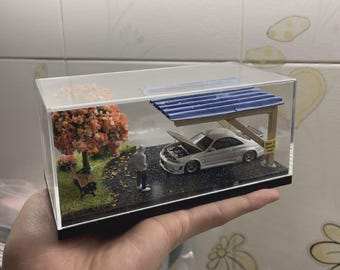 Può includere: Un diorama in miniatura all'interno di una teca in acrilico trasparente con un'auto sportiva bianca dal cofano aperto sotto un tetto blu ondulato. Una piccola figura si trova su un sentiero di ghiaia vicino a una panchina e un albero con foglie arancioni.