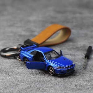 Puede incluir: Un coche de modelo fundido a presión azul con una puerta abierta, sujeto a un llavero con una correa de cuero marrón. El coche tiene ruedas doradas y un interior negro. También se ve un pequeño destornillador negro.