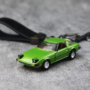 May include: A lime green die-cast sports car keychain featuring pop-up headlights, a detailed interior, and silver wheels. The miniature vehicle is attached to a black leather strap with a metal ring, showcasing a classic coupe design.