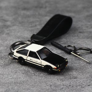 May include: A black and white toy car keychain with a black leather strap. The car has a black hood and roof, with white body panels. The keychain also includes a black metal ring and a small tool.