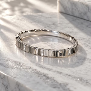 May include: A silver-toned hinged bangle bracelet featuring a series of rectangular metallic links. The design alternates between smooth, polished silver-colored segments and smaller, inset links encrusted with clear, sparkling stones.