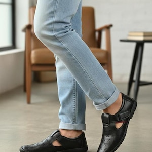 Puede incluir: Sandalias de cuero negro con diseño perforado y correa ajustable. Las sandalias tienen punta redonda y tacón bajo. La persona lleva vaqueros azul claro.