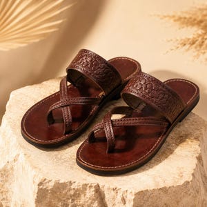 May include: A pair of dark brown leather sandals featuring a wide, embossed floral strap across the instep and crisscrossing straps that loop around the big toe. The sandals have a flat sole with visible stitching along the edges of the footbed.