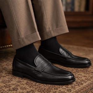May include: A pair of black leather penny loafers with a smooth finish, a rounded toe, and a classic strap across the vamp. The shoes are worn with solid black ribbed socks and tan trousers that feature a cuffed hem at the bottom of the legs.