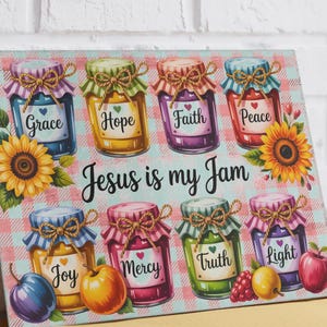 Könnte beinhalten: Ein rechteckiges Glasschneidebrett mit hellblau-rosa kariertem Hintergrund und acht bunten Gläsern, die mit Grace, Hope, Faith, Peace, Joy, Mercy, Truth und Light beschriftet sind. Der Text Jesus is my Jam ist mittig mit Sonnenblumen platziert.