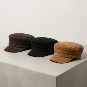 Può includere: Tre berretti in stile militare nei colori marrone scuro, nero e marrone chiaro. Ogni berretto presenta una parte superiore piatta, una visiera corta e un cinturino orizzontale decorativo alla base, fissato con piccoli bottoni metallici.