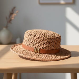 May include: A tan woven straw hat with a flat top and a wide brim. The hat features a smooth brown leather band encircling the base of the crown, accented with four vertical metallic gold bars positioned at the front centre of the hat.