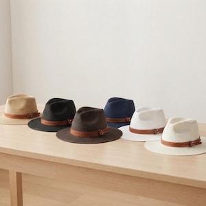 Op de afbeelding: Een collectie van zes fedora hoeden in beige, zwart, donkerbruin, marineblauw en wit. Elke hoed heeft een geweven textuur en een bruine leren band met een klein metalen gespdetail rond de kroon, gerangschikt in een rij op een vlak oppervlak.